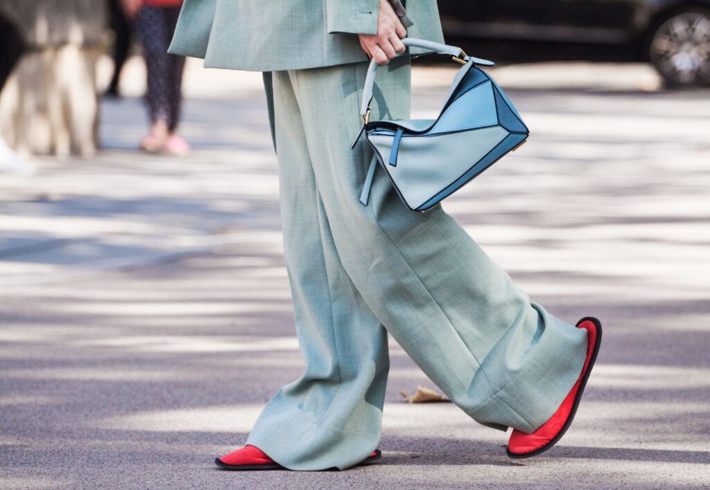 Imagen de la parte inferior del cuerpo de una mujer vestida con traje pantalón ancho en tono verde pastel apagado y un bolso de cuero celeste. Remata el look con unas zapatillas propias de las que ofrecen los hoteles como amenities