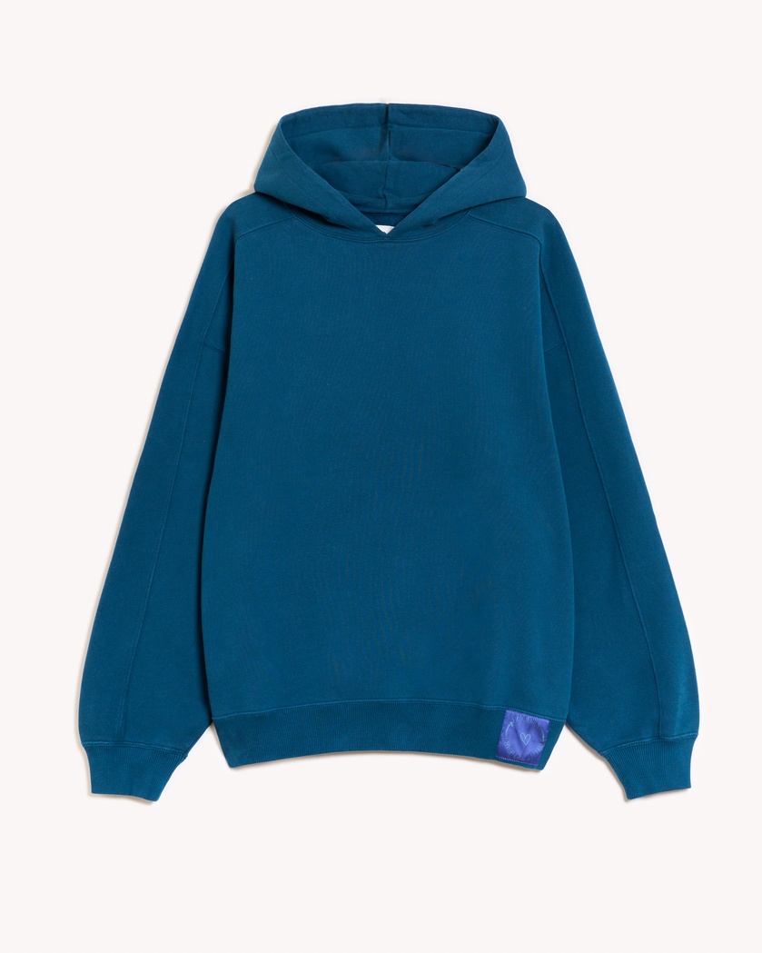 Imagen de la parte delantera de la hoodie modelo Cistierna con capucha en color azul turquesa oscuro. En la parte izquierda del elástico inferior de la sudadera lleva cosido en tela el logo de hoodies with heart. Modelo unisex de corte holgado y hombro caído.