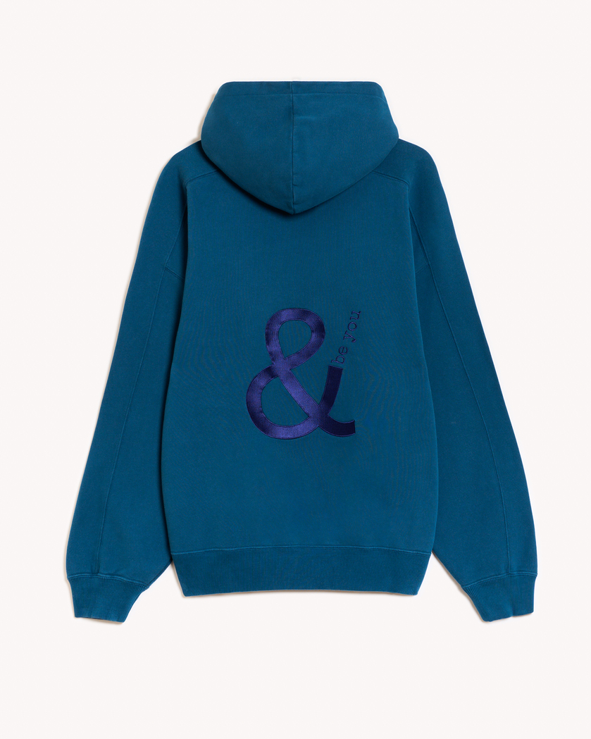 Imagen de la parte trasera de la hoodie modelo Cistierna con capucha en color azul turquesa oscuro con el logo de and be you brand bordado en color azul marino. Modelo unisex de corte holgado y hombro caído.
