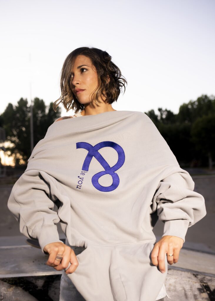 Imagen que muestra a una modelo femenina con la hoodie Cistierna con capucha en color gris claro. Solo se le ve el tronco superior y la sudadera se ve por la parte trasera con el logo de and be you brand bordado en color azul marino porque la modelo se dispone a ponérsela. No está mirando a la cámara de frente sino que está mirando a su derecha y en el fondo se aprecian unos árboles con cierta dificultad porque están difuminados.