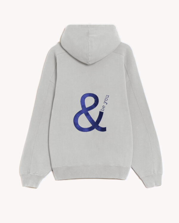 Imagen de la parte trasera de la hoodie modelo Cistierna con capucha en color gris claro con el logo de and be you brand bordado en azul marino. Modelo unisex de corte holgado y hombro caído.