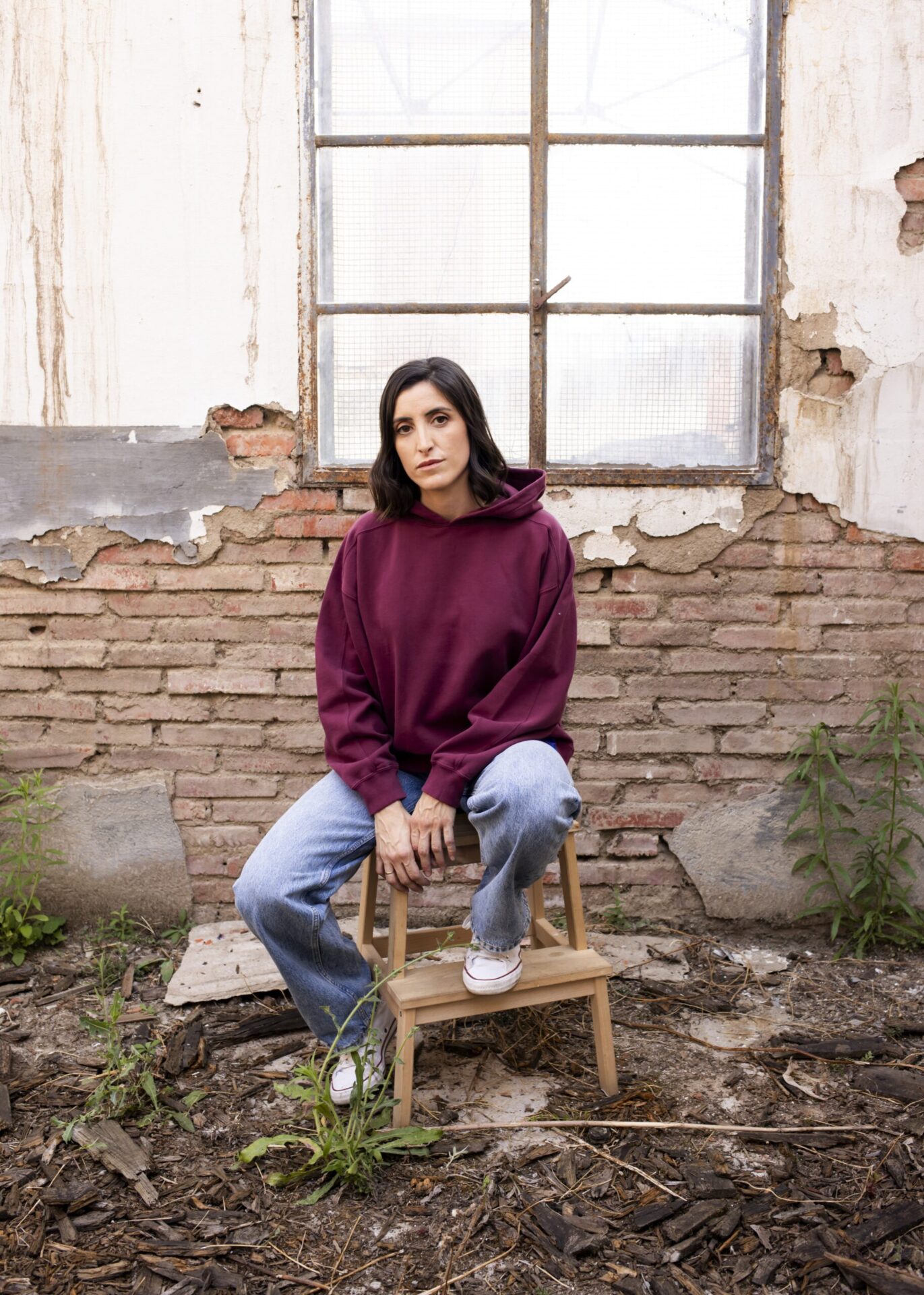 Imagen que muestra a una modelo femenina con la hoodie Cistierna con capucha en color granate. Se encuentra sentada sobre un taburete de madera con unos vaqueros azul claro y unas bambas en color blanco. De fondo se aprecia una fachada desconchada y una ventana desvencijada.