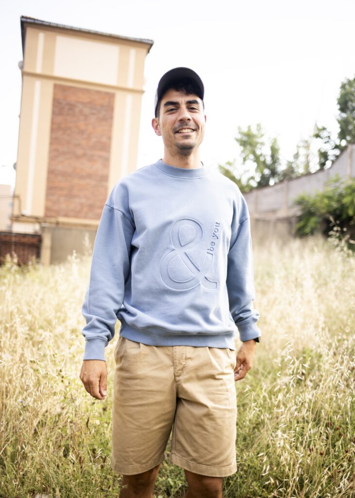 Imagen que muestra a un modelo masculino con la sudadera Adrados sin capucha en color azul claro añil con el logo de and be you brand en bajo relieve. Se encuentra de pie sonriendo con una bermuda beige y una gorra gris oscuro. Está en el campo entre la maleza y de fondo se aprecia una torre de un edificio.