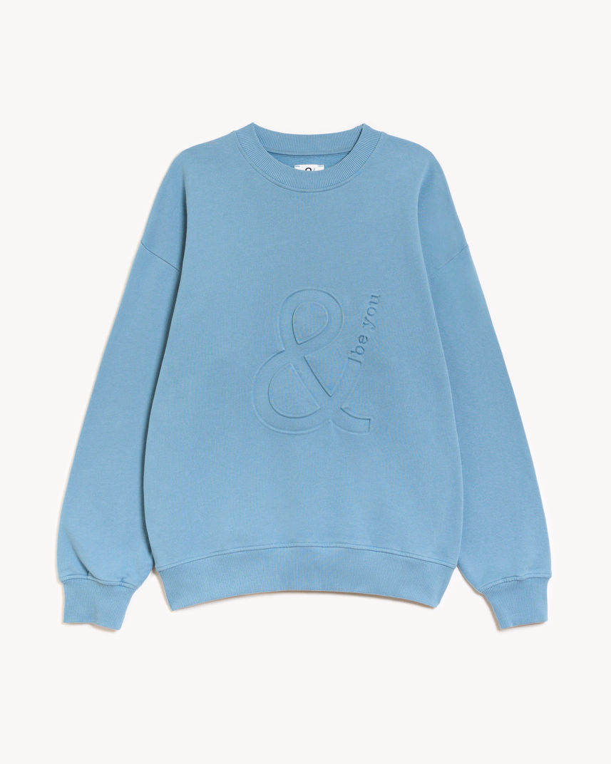 Imagen de la parte delantera de la sudadera modelo Adrados sin capucha en color azul claro añil con el logo de and be you brand en bajo relieve. Modelo unisex de corte holgado y hombro caído.