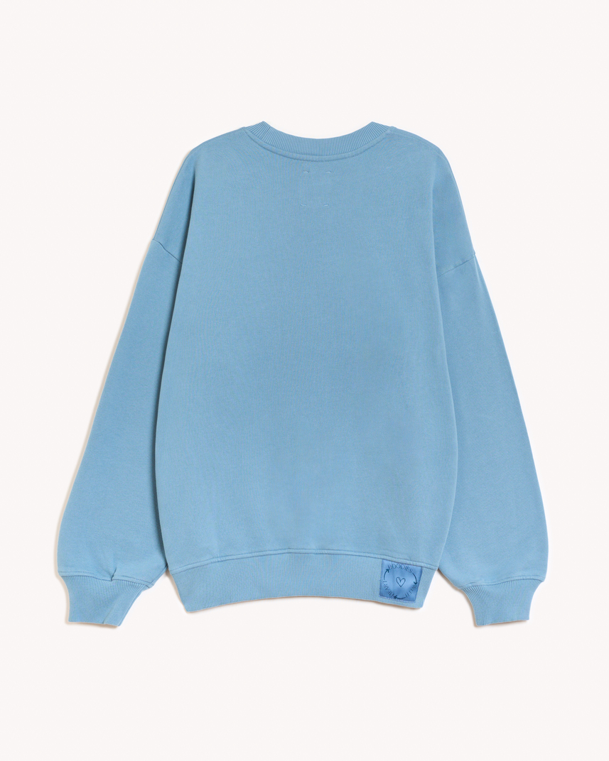 Imagen de la parte trasera de la sudadera modelo Adrados sin capucha en color azul claro añil. En la parte derecha del elástico inferior de la sudadera lleva cosido en tela el logo de hoodies with heart. Modelo unisex de corte holgado y hombro caído.