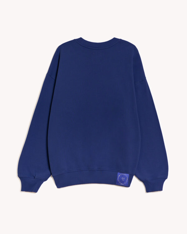 Imagen de la parte trasera de la sudadera modelo Adrados sin capucha en color azul intenso. En la parte derecha del elástico inferior de la sudadera lleva cosido en tela el logo de hoodies with heart. Modelo unisex de corte holgado y hombro caído.